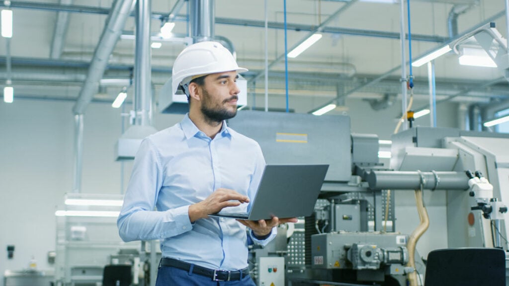 Um homem de pé, usando camisa social, calça jeans e capacete, segurando um laptop, em frente às máquinas, avalia a automação de processos industriais