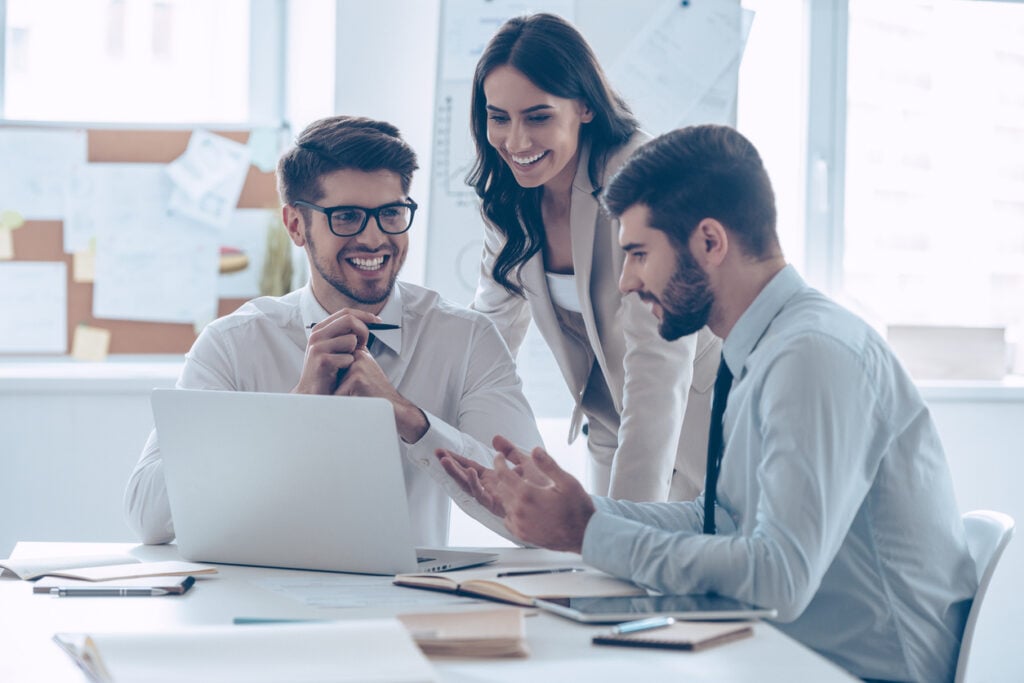 dois homens e uma mulher sorriem, em volta da mesa, com computador e documentos, em um bom ambiente de trabalho