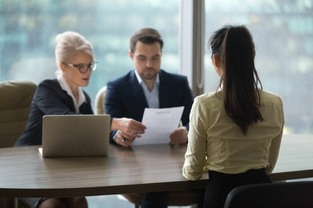 Uma mulher de costas, com os cabelos presos, participa de uma entrevista de emprego conduzida por uma mulher de cabelos brancos e um homem de barba